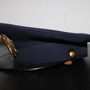 以下が含まれることがあります： 黒い縁と金色のエンブレムが付いた濃紺の帽子。帽子は、わずかに湾曲したつばを持つ構造化された形状をしています。帽子の側面には金のボタンが付いています。帽子はウールのような素材で作られているようです。