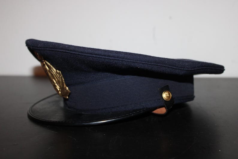 以下が含まれることがあります： 黒い縁と金色のエンブレムが付いた濃紺の帽子。帽子は、わずかに湾曲したつばを持つ構造化された形状をしています。帽子の側面には金のボタンが付いています。帽子はウールのような素材で作られているようです。