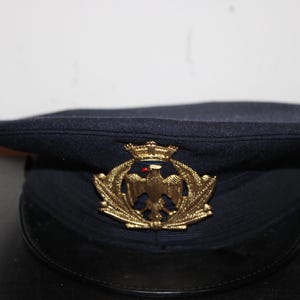以下が含まれることがあります： 黒いひさしが付いた濃紺のミリタリーハット。帽子には、王冠をかぶったワシの金色のエンブレムが付いています。この帽子はおそらくヴィンテージまたはコレクターズアイテムです。