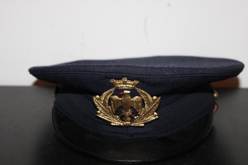 以下が含まれることがあります： 黒いひさしが付いた濃紺のミリタリーハット。帽子には、王冠をかぶったワシの金色のエンブレムが付いています。この帽子はおそらくヴィンテージまたはコレクターズアイテムです。