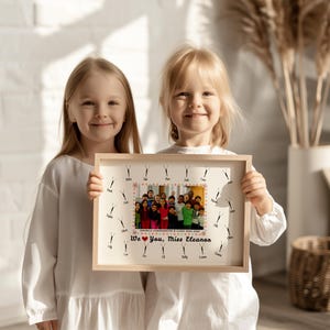 May include: Two young girls hold a framed photo with a group of children. The frame is light wood, and the photo is surrounded by decorative elements and names. The text on the frame reads "We ❤️ You, Miss Eleanor" and "Family Kindergarten & Class 2024-2025".