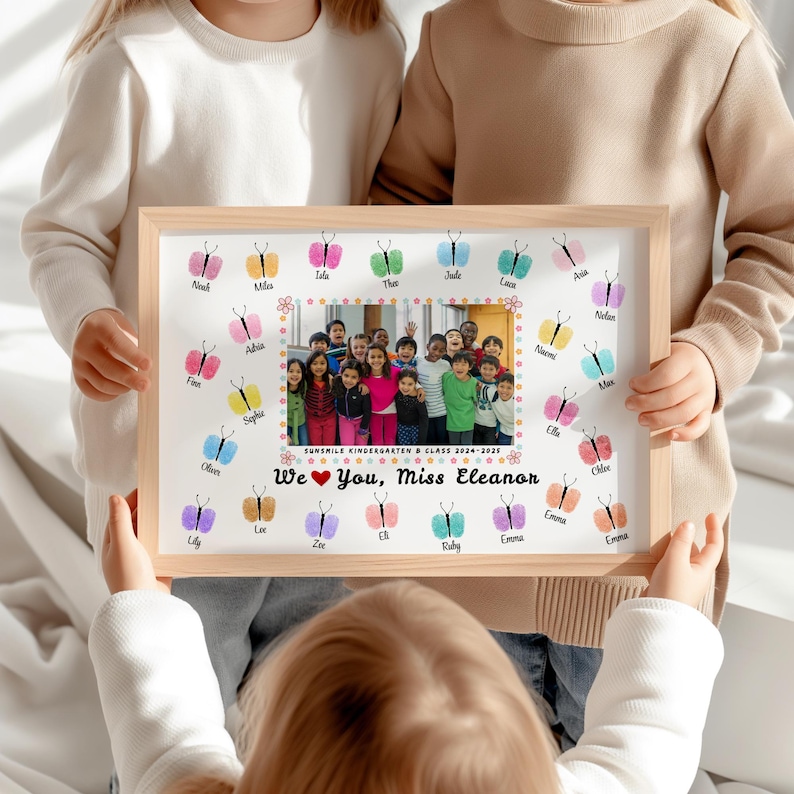 May include: A framed print featuring a class photo surrounded by colorful butterfly prints. The text "We ❤️ You, Miss Eleanor" is displayed below the photo. The frame is held by children.