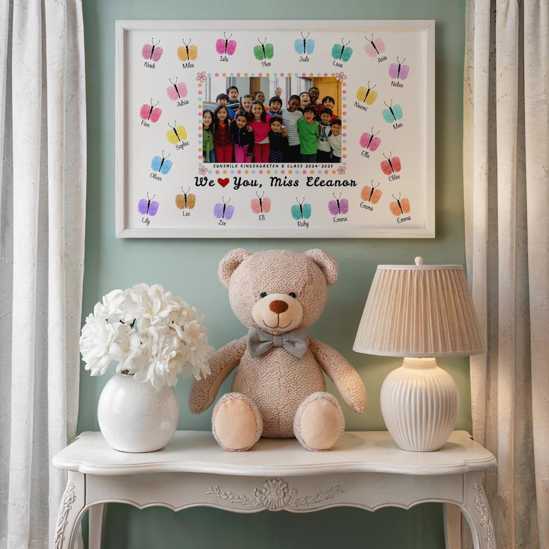 May include: A framed class photo with butterfly thumbprints and the text "We ❤️ You, Miss Eleanor." A plush teddy bear with a bow tie, a white vase with flowers, and a lamp with a beige shade sit on a white table.