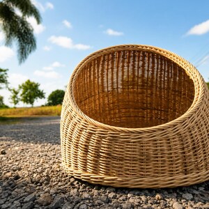 May include: A woven wicker basket with a rounded shape and open top. The basket is light brown and sits on a gravel surface, with a blue sky and trees in the background. The basket is designed for storage.