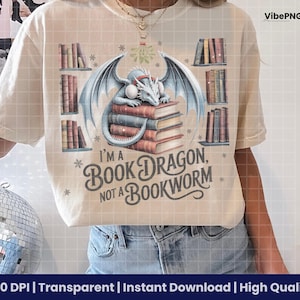 May include: Cream-colored t-shirt featuring a dragon atop a stack of books, with the text "I'M A BOOK DRAGON, NOT A BOOKWORM." The design includes illustrations of bookshelves and snowflakes. The shirt is a comfortable, casual wear.