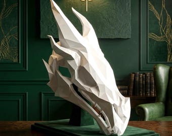May include: A white, low-poly geometric dragon skull sculpture mounted on a dark green rectangular base. The sculpture features sharp, angular planes and pointed horns, creating a stylized, faceted representation of a dragon head in profile.