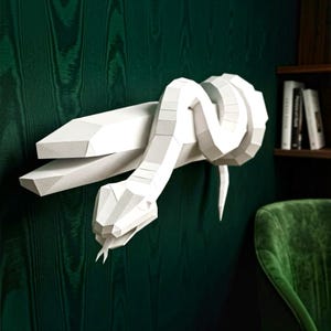 May include: A white, low-poly papercraft sculpture of a snake coiled around a large, faceted crystal-like structure. The snake features a geometric, angular design with a visible tongue, mounted as a wall decoration with a matte white finish.