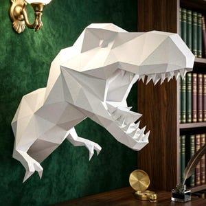 Può includere: Una scultura da parete a forma di testa di dinosauro T-Rex geometrica bianca, caratterizzata da un design sfaccettato low-poly con bocca aperta che mostra denti affilati e due piccoli artigli anteriori che sporgono dal collo, in carta bianca opaca.