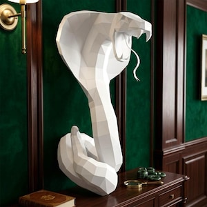 Puede incluir: Una escultura de pared geométrica de bajo poligonaje en color blanco que representa una cobra con la capucha desplegada y la boca abierta, mostrando colmillos y una lengua fina y curva. La pieza presenta facetas angulares y acabado mate.