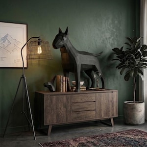 May include: A dark brown wooden sideboard with three central drawers and two side cabinets. On top sits a large black geometric dog sculpture, a stack of vintage books, a small white bowl, and a magnifying glass on a stand.