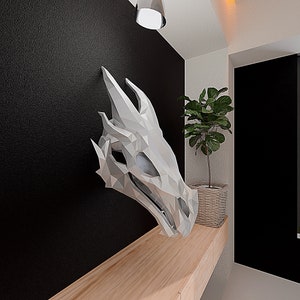 May include: A white origami dragon head sculpture mounted on a wooden shelf. The dragon head is made of paper and has a geometric design. The shelf is light brown and has a black wall behind it.