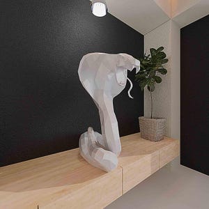 May include: A white, geometric cobra sculpture with its hood raised and tongue extended. The artwork is displayed on a light wood shelf against a black wall. A small potted plant in a woven basket sits nearby.