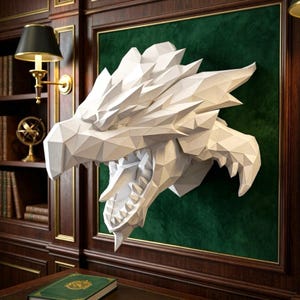 May include: A white, low-poly geometric dragon head sculpture mounted on a dark green velvet panel. The sculpture features sharp, angular facets, a wide-open mouth with visible teeth, and a pointed snout, creating a structured, three-dimensional form.