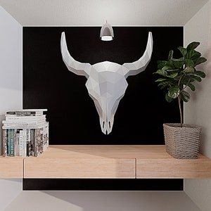 May include: A white origami bull skull hanging on a black wall with a small light fixture above it. A potted plant with green leaves sits on a light wood shelf below the skull.