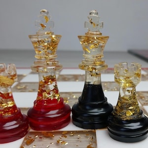 Puede incluir: Juego de ajedrez con piezas de resina transparente y detalles de pan de oro. Los reyes y las torres son transparentes con copos de oro, y las bases son rojas y negras. El tablero de ajedrez tiene un patrón de cuadros blancos y dorados.