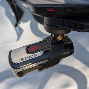 May include: A silver and black bicycle light with the word "SIGMA" printed on it. The light has a red button with a white circle and a black line through it. The light is attached to a black mount with a black strap.