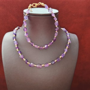 May include: A set of two amethyst necklaces with gold accents. The necklace is made of small, faceted amethyst beads and is strung on a gold chain. The shorter necklace is a bracelet.