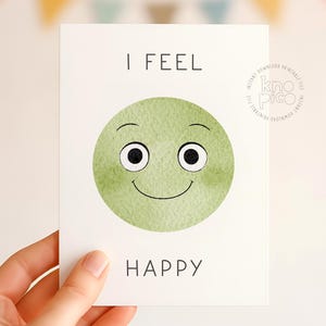 May include: A white card with the words "I FEEL HAPPY" in black text. The card features a green watercolor smiley face with black eyes and a curved smile. The card is held by a hand.