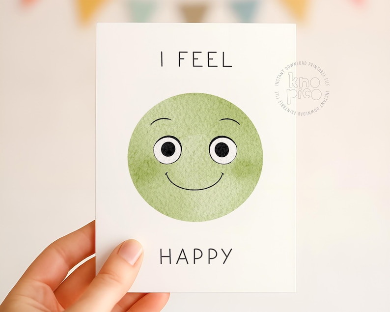May include: A white card with the words "I FEEL HAPPY" in black text. The card features a green watercolor smiley face with black eyes and a curved smile. The card is held by a hand.