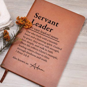 May include: A brown leather-bound journal with the words "Servant Leader" printed in black. The journal includes a definition and inspirational text. A sprig of orange flowers rests on the journal's cover.