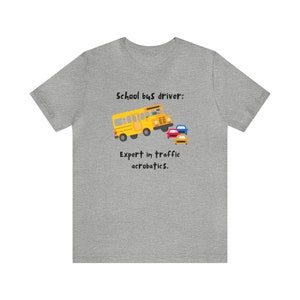 Puede incluir: Una camiseta gris con el texto "School bus driver: Expert in traffic acrobatics." y una imagen de dibujos animados de un autobús escolar amarillo que se cuela entre tres coches.