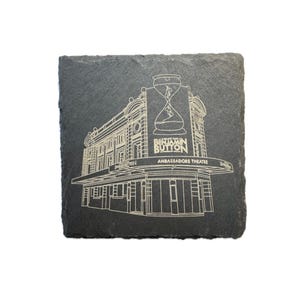 May include: A black slate coaster with a gold engraved illustration of the Ambassadors Theatre in London, featuring the text "The Curious Incident of the Dog in the Night-Time" and "Ambassadors Theatre".