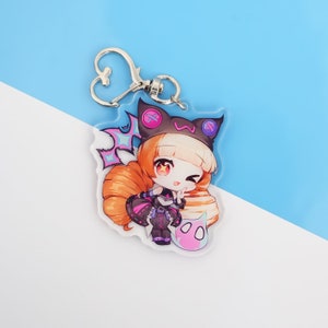 Pu&ograve; includere: Un portachiavi con un personaggio in stile anime colorato con un design di gatto rosa e viola. Il personaggio ha i capelli arancioni, un vestito bianco e nero e un accessorio per la testa a forma di gatto rosa. Il portachiavi &egrave; attaccato a un anello di metallo argentato.