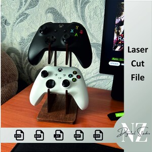 May include: Laser cut file for a two-tiered stand to hold two Xbox controllers. The stand is made of wood and has a dark brown finish. The controllers are black and white. The file is available in SVG, DXF, AI, PDF, and CDR formats.