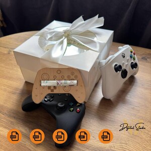 May include: A white gift box with a white satin ribbon and bow. The box is open and contains a wooden controller stand with a rolled up bill inside. A white and black video game controller is on the table next to the box.