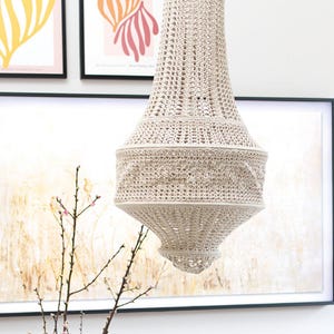 Pode incluir: Um candelabro de crochê creme pendura do teto. A luminária tem formato de sino com um padrão decorativo no meio. O fundo inclui impressões de arte emolduradas e um galho de galhos floridos.