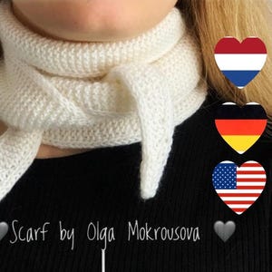 May include: A white knitted scarf with a textured stitch pattern wrapped around the neck. The scarf features pointed ends and is worn over a black top. The text Scarf by Olga Mokrousova is visible at the bottom along with three heart-shaped flag icons.