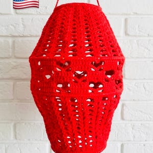 May include: A handmade, red crochet lantern with a unique openwork design. The lantern is suspended by a matching red cord, showcasing a textured, open weave pattern. The decorative piece is designed to hold a light source.