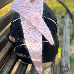 Può includere: Una sciarpa lavorata a maglia di colore rosa chiaro e dall'aspetto soffice, drappeggiata su uno zaino nero trapuntato. La sciarpa presenta una trama a maglia larga con un aspetto morbido simile al mohair. Lo zaino ha un motivo a rombi e cerniere argentate.