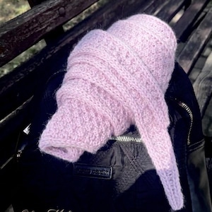 May include: A soft, light pink knitted scarf with a textured, ribbed pattern, coiled into a rounded shape. The scarf features a fuzzy, wool-like texture and is draped over a black backpack with a visible silver zipper and a small rectangular logo tag.