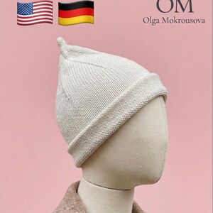 Puede incluir: Un gorro de punto color crema con una parte superior puntiaguda y un borde doblado. El gorro se muestra sobre una cabeza de maniqu&iacute;. El texto en la esquina superior derecha dice OM Olga Mokrousova, junto a las banderas de EE. UU. y Alemania.