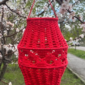 May include: A bright red crocheted lantern featuring an intricate openwork pattern with a wide, rounded body and a narrow top opening. The lantern includes a thin red yarn handle attached to the rim, allowing it to be suspended from a branch.