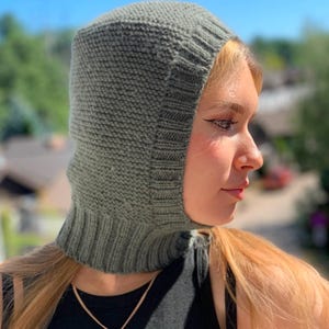 May include: A gray knitted balaclava with a long neck covering. The balaclava is worn by a person with long blonde hair and a gold necklace with a small pendant.