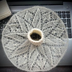 Könnte beinhalten: Ein kreisförmiges, cremeweißes Strickstück mit einem zarten, durchbrochenen Spitzenmuster und einem zentralen Rippenkragen. Das Textil hat eine flauschige, mohairartige Textur und wird flach ausgebreitet gezeigt, wobei die komplexen, floralen Motive sichtbar sind.