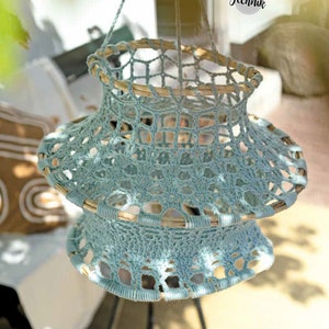 Peut inclure: Un abat-jour en crochet bleu clair avec un anneau en bois en haut et en bas. L'abat-jour est suspendu à un plafond blanc.