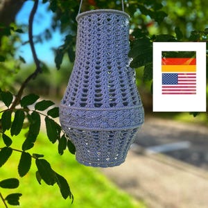 Könnte beinhalten: Eine hellblaue, handgehäkelte Laterne, die an einem Ast hängt. Die Laterne hat eine zylindrische Form mit einem durchbrochenen Design und einem dekorativen Rand. Der Hintergrund zeigt grünes Laub und einen verschwommenen Weg. Ein kleines Bild der deutschen und amerikanischen Flaggen ist im Hintergrund.