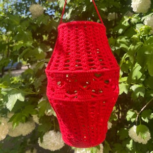 May include: A vibrant red crochet lantern, featuring an openwork pattern. The lantern is cylindrical with a matching red yarn handle. The background shows green leaves and white flowers, suggesting an outdoor setting.