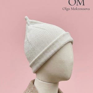 May include: A cream-colored knit beanie with a pointed top and a wide folded brim. The hat is displayed on a mannequin head. The text OM Olga Mokrousova is visible in the upper right corner of the image, set against a solid light pink background.