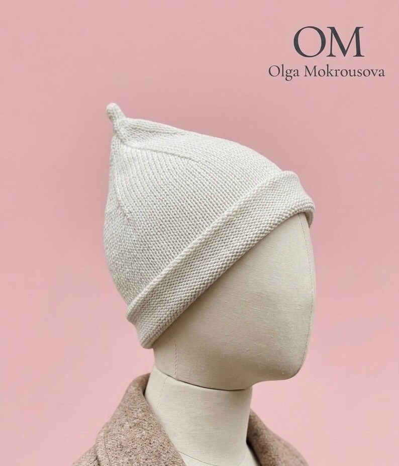 Puede incluir: Un gorro de punto color crema con la parte superior puntiaguda y un ala ancha doblada. El gorro se muestra sobre una cabeza de maniqu&iacute;. El texto OM Olga Mokrousova es visible en la esquina superior derecha sobre un fondo rosa claro.
