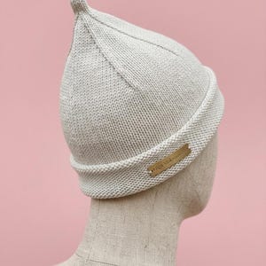 Puede incluir: Un gorro de punto de color beige claro con la parte superior puntiaguda y un borde doblado. Una peque&ntilde;a etiqueta met&aacute;lica rectangular est&aacute; fijada en el lateral del borde con el texto grabado Olga Mokrousova.