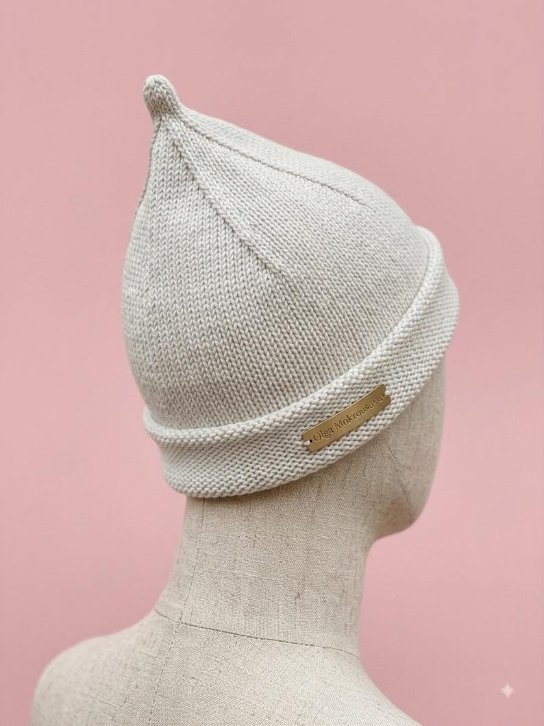 Puede incluir: Un gorro de punto de color beige claro con la parte superior puntiaguda y un borde doblado. Una peque&ntilde;a etiqueta met&aacute;lica rectangular est&aacute; fijada en el lateral del borde con el texto grabado Olga Mokrousova.