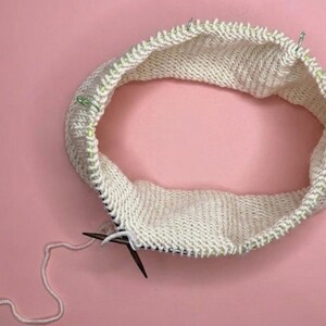Puede incluir: Un proyecto de tejido circular en progreso con hilo color blanco hueso en una aguja circular. La pieza est&aacute; tejida en punto jersey con varios marcadores de puntos de pl&aacute;stico verde sujetos a lo largo del borde superior del tejido.