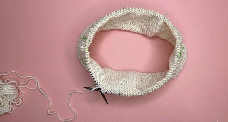Puede incluir: Un proyecto de tejido circular en progreso con hilo color blanco hueso en una aguja circular. La pieza est&aacute; tejida en punto jersey con varios marcadores de puntos de pl&aacute;stico verde sujetos a lo largo del borde superior del tejido.