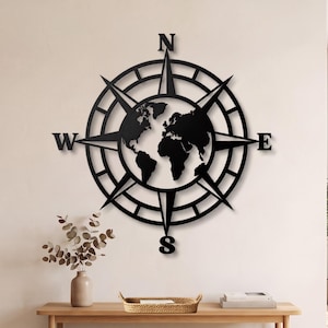 Può includere: Un'opera d'arte da parete in metallo nero caratterizzata da un design a rosa dei venti circolare con una sagoma della mappa del mondo al centro. La bussola include le lettere cardinali N, S, E e O posizionate nei punti alto, basso, destra e sinistra.