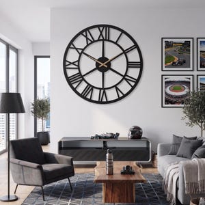May include: A large, black, round wall clock with Roman numerals and gold-coloured hands. The clock is mounted on a white wall in a modern living room setting. The room includes a grey armchair, a wooden coffee table, and framed pictures.