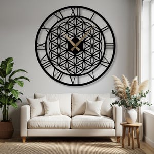 Puede incluir: Un gran reloj de pared decorativo negro con números romanos y un diseño geométrico de la flor de la vida. Las manecillas del reloj son doradas. El reloj está montado en una pared blanca sobre un sofá blanco.